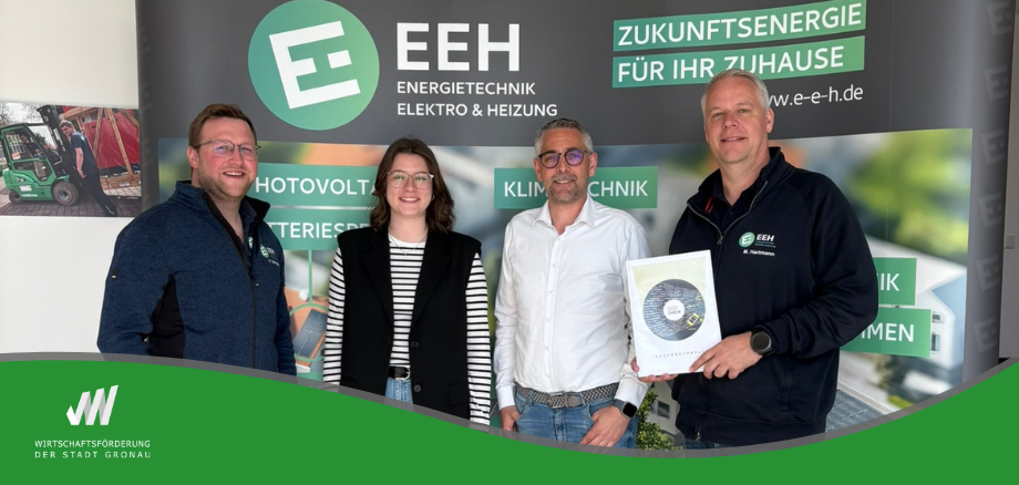 Vier Personen stehen gemeinsam vor einem Banner der Firma „EEH Energietechnik Elektro & Heizung“ und blicken in die Kamera. Rechts hält ein Mitarbeiter eine gerahmte Auszeichnung in den Händen. Im Hintergrund sind Begriffe wie „Photovoltaik“, „Klimatechnik“ und „Zukunftsenergie für Ihr Zuhause“ zu sehen. Am unteren Bildrand befindet sich das Logo der Wirtschaftsförderung der Stadt Gronau. 