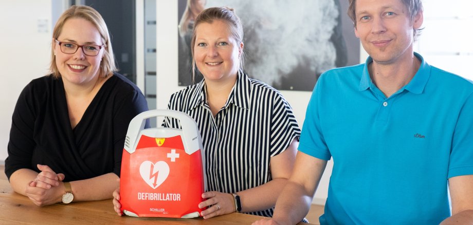 Auf dem Bild sehen Sie links 2 Frauen und rechts einen Mann sitzen. In der Mitte des Bildes steht ein Defibrillator.