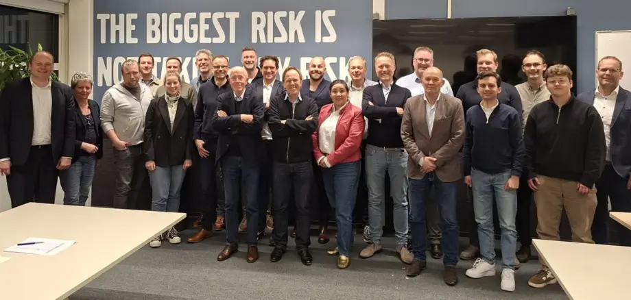 Gruppenfoto mit mehreren Personen in einem Besprechungsraum. Die Teilnehmenden stehen nebeneinander und blicken in die Kamera. Im Hintergrund befindet sich ein großer Schriftzug an der Wand.