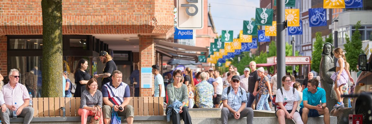 Belebte Fußgängerzone mit zahlreichen Menschen, Sitzgelegenheiten und bunten Fahnen über der Straße. Im Vordergrund sitzen mehrere Personen auf Bänken, rechts ist eine Statue zu sehen.