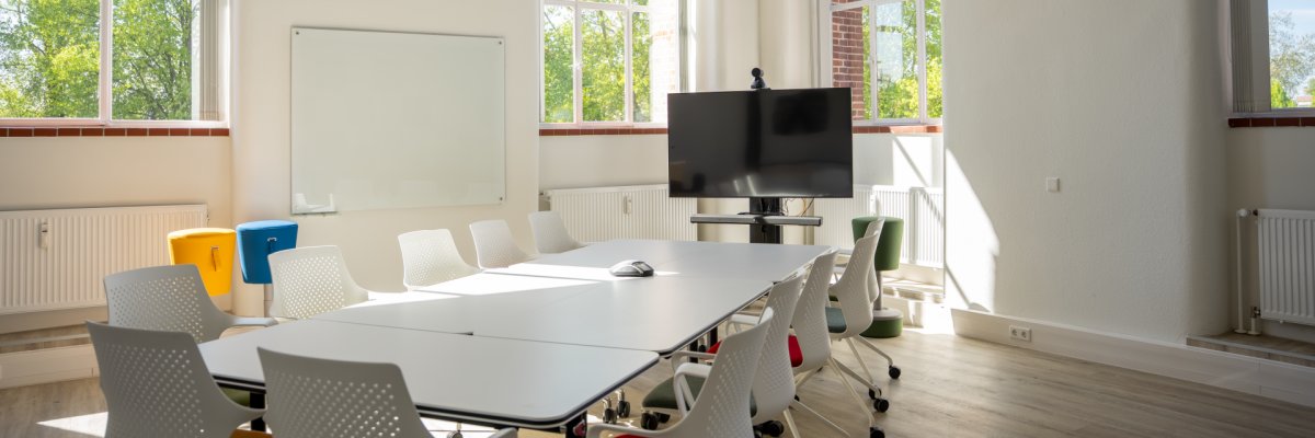 Heller Besprechungsraum mit großen Fenstern, mehreren weißen Stühlen und einem langen Tisch. Im Hintergrund steht ein großer Bildschirm für Präsentationen oder Videokonferenzen.
