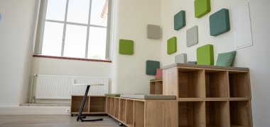 Heller Innenraum mit großem Fenster, Sitzmöglichkeiten aus Holz und farbigen Wandpaneelen in Grün- und Grautönen. Der Raum wirkt modern und freundlich gestaltet.