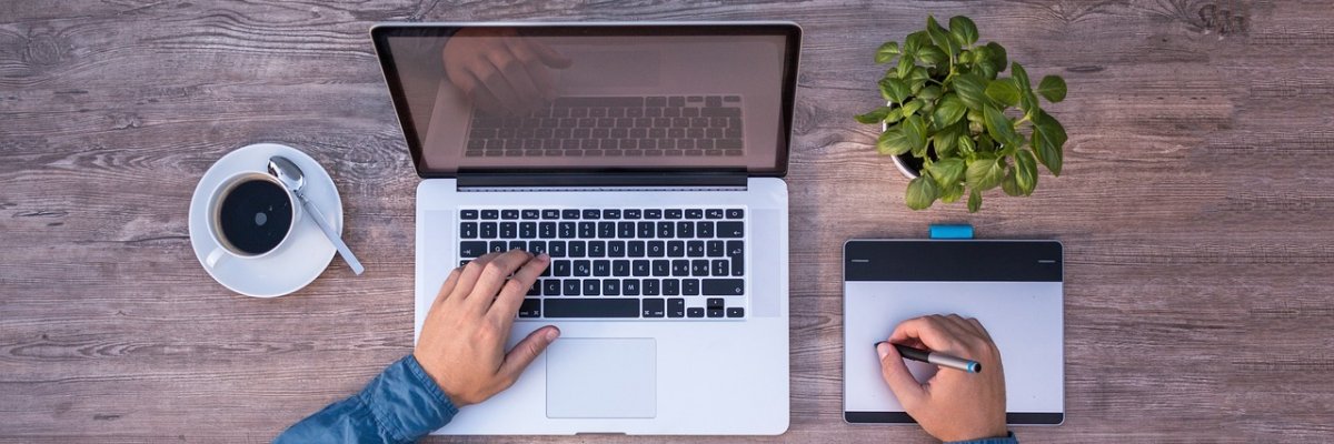 Blick von oben auf einen Schreibtisch mit Laptop, Grafiktablett, Kaffeetasse und kleiner Pflanze. Eine Person arbeitet gleichzeitig an Tastatur und Grafiktablett.