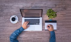 Blick von oben auf einen Schreibtisch mit Laptop, Grafiktablett, Kaffeetasse und kleiner Pflanze. Eine Person arbeitet gleichzeitig an Tastatur und Grafiktablett.
