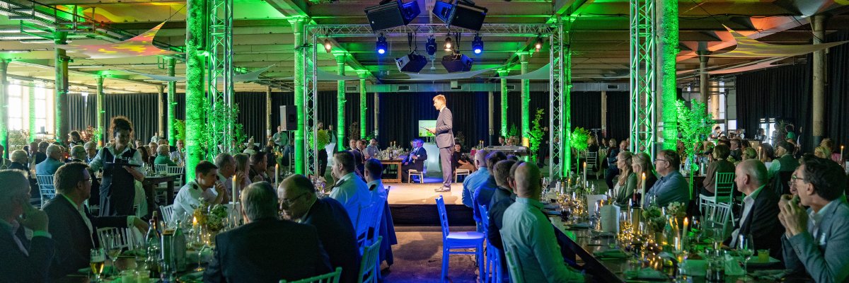 Das Bild zeigt eine Abendveranstaltung in einer Halle mit vielen Gästen an gedeckten Tischen. In der Mitte steht eine Person auf einer Bühne und spricht vor dem Publikum. Der Raum ist mit grüner Beleuchtung ausgeleuchtet.