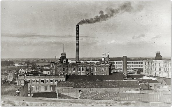 Historische Schwarz-Weiß-Aufnahme eines Industrieareals mit mehreren Fabrikgebäuden und einem hohen Schornstein, aus dem Rauch aufsteigt. Im Hintergrund sind weitere Gebäude und die umliegende Landschaft zu erkennen.
